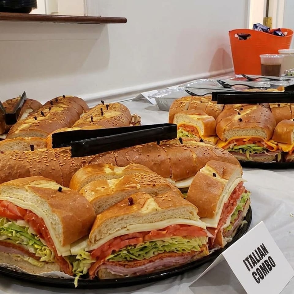 Buffet table displaying assorted Italian combo sandwiches with deli meats, lettuce, tomatoes, and sliced rolls.